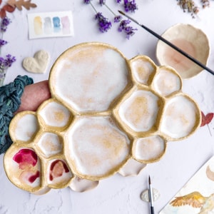 Könnte beinhalten: Eine keramische Aquarellpalette in Wolkenform mit mehreren unregelmäßigen Vertiefungen. Die Palette hat einen strukturierten, goldbraunen Rand und eine glänzende, cremeweiße Innenfläche. Zwei der kleineren Vertiefungen enthalten Spuren von rosa und roter Farbe.