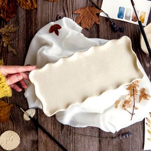Könnte beinhalten: Ein cremefarbenes, rechteckiges Keramik-Serviertablett mit gewelltem Rand. Das Tablett liegt auf einem weißen Tuch, umgeben von Herbstblättern, Pinseln und Aquarellmustern. Eine Hand in einem gelben Pullover berührt das Tablett.