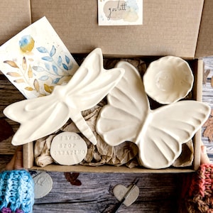 May include: A gift box containing a cream-coloured ceramic dragonfly dish, a butterfly dish, a small round bowl, and a circular tag with the text NEVER STOP CREATING. Also included is a watercolour illustration of leaves and a small branded card.