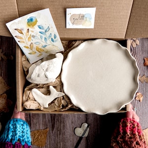 May include: A cardboard box containing handmade ceramics. It features a large, off-white plate with a wavy edge, a small leaf-shaped bowl, and a bird-shaped figurine. A watercolour painting and a small card with the word "golett" are also visible.