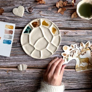 Peut inclure: Un espace de travail d'artiste céramiste avec une palette de peinture en céramique blanche contenant diverses couleurs, une carte d'échantillons d'aquarelle et une illustration de cerf en cours de peinture. La scène est sur une surface en bois avec des feuilles d'automne.