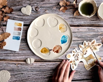 May include: A cream-colored ceramic artist's palette with eight wells, some filled with colorful paint. A watercolor swatch card and a deer illustration are also present. The scene is set on a rustic wooden surface.