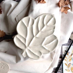 Peut inclure: Une palette en céramique en forme de feuille, de couleur crème, avec des motifs de veines détaillés. La palette repose sur un tissu blanc, avec des feuilles séchées et un pinceau à proximité. Une aquarelle est également visible.