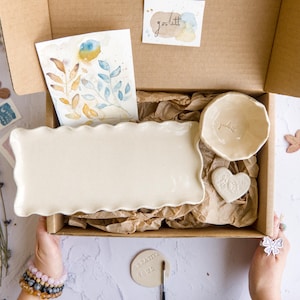 Puede incluir: Una caja de regalo de cartón que contiene una bandeja rectangular de cerámica color crema con bordes ondulados, un pequeño cuenco de cerámica a juego con el texto "golitt", una pieza de cerámica en forma de corazón y una acuarela de hojas azules y doradas.