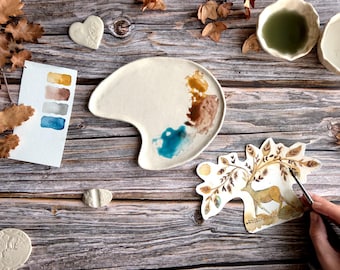 May include: A ceramic artist's workspace featuring a palette with paint, watercolor swatches, and a deer illustration. The palette has brown, blue, and tan paint. The watercolor swatches show shades of brown, gray, and blue. The deer illustration is in shades of brown and tan.