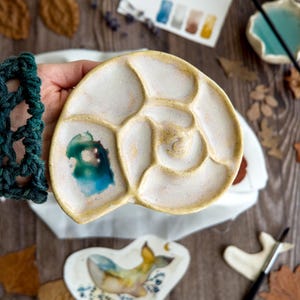 Könnte beinhalten: Eine Keramik-Farbpalette in Form eines Nautilus-Gehäuses mit glänzend cremeweißer Oberfläche und gelblichen Rändern. Die Palette verfügt über mehrere geschwungene Vertiefungen zum Mischen von Farben, wobei ein kräftiges Türkis in einem der Felder zu sehen ist.