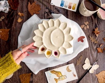 May include: A cream-colored ceramic artist's palette in a sunflower shape, with a central well and several color wells filled with paint. The palette rests on a white cloth, surrounded by autumn leaves and art supplies.