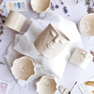 Può includere: Una collezione di vasi e ciotole in ceramica fatti a mano con volti, in una palette di colori neutri. I vasi e le ciotole hanno superfici strutturate e design di volti unici. L'allestimento comprende acquerelli e fiori secchi.