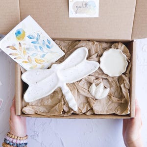 Peut inclure: Un coffret cadeau contenant un plat de service en céramique blanche en forme de libellule, une petite assiette ronde et un plat en forme de feuille. La boîte comprend également une aquarelle d'éléments floraux bleus et jaunes. Les articles sont disposés sur du papier d'emballage brun.