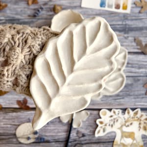 Könnte beinhalten: Eine cremefarbene Keramikschale in Blattform mit detaillierten Adern. Die Schale wird von einer Person gehalten, die einen beigefarbenen Strickärmel trägt. Dekorative Blätter und eine Aquarellpalette befinden sich im Hintergrund.