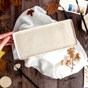 May include: A rectangular, off-white ceramic tray is held over a white cloth. The tray is surrounded by autumn leaves, watercolor paints, and a small bowl. A yellow sweater sleeve is visible in the corner.