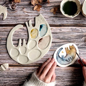 Può includere: Una tavolozza per pittura in ceramica color crema a forma di gatto con diversi pozzetti pieni di colori. Un acquerello di un gatto è accanto alla tavolozza. La tavolozza è su una superficie di legno.