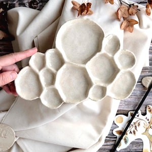 Può includere: Una tavolozza per pittura in ceramica bianca fatta a mano con più pozzetti a forma di nuvola. La tavolozza è esposta su un panno color crema, con un pennello ed elementi decorativi nelle vicinanze. Ideale per gli artisti.