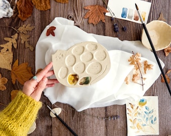May include: A speckled, off-white ceramic paint palette with multiple circular wells, partially filled with colorful paint. The palette sits on a white cloth, surrounded by autumn leaves, watercolor paintings, and paintbrushes. The palette is held by a person wearing a yellow sweater.