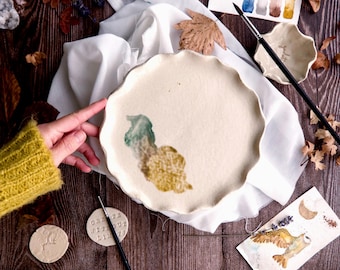 May include: A cream-colored ceramic plate with a scalloped edge, featuring abstract watercolor designs in shades of green and gold. The plate rests on a white cloth, with paintbrushes and a watercolor palette nearby. The words "CREATE WITHOUT LIMITS" are stamped on a small ceramic disc.