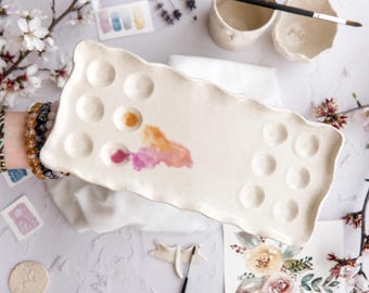 May include: A cream-colored ceramic paint palette with 14 circular wells and a wavy edge. The palette has a watercolor stain of pink, orange, and yellow. The image also shows paintbrushes, small ceramic bowls, and watercolor paper.