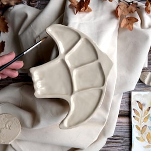 May include: A cream-colored ceramic artist's palette with multiple paint wells. A hand holds a paintbrush over the palette. A small, round, embossed ceramic piece and a watercolor painting are also visible.