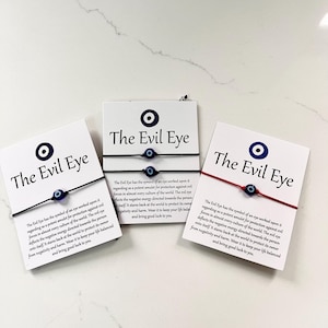 May include: Three Evil Eye bracelets on white cards. Each card has the text "The Evil Eye" and a description. The bracelets are black and red string with a blue and white eye charm. The cards are on a white surface.