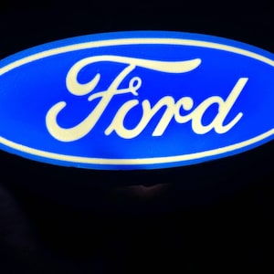 May include: A blue oval logo featuring the word "Ford" in white script. The logo has a white border and is set against a black background. The image is well-lit, highlighting the logo's design.