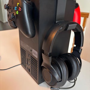 May include: A black Xbox Series X console with a black controller and black headphones attached to the side. The console is sitting on a white surface.