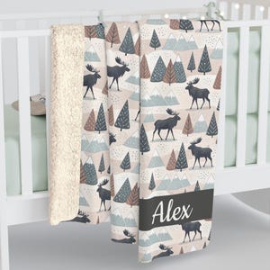 Peut inclure: Une couverture douce à motifs sur le thème de la forêt, avec des élans, des arbres et des montagnes dans des tons de gris, de marron et de bleu. La couverture a un envers en peluche crème et une bande gris foncé avec le nom "Alex" en blanc.