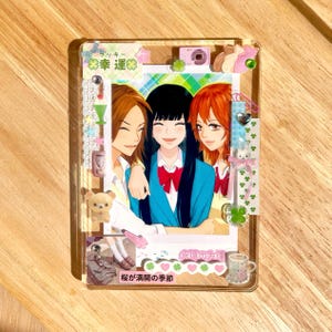 May include: A clear acrylic photo holder featuring a colorful anime-style illustration of three smiling women. The holder is decorated with small charms, including a camera, a bear, and a four-leaf clover. Japanese text is visible.