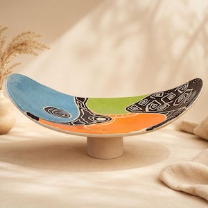 Könnte beinhalten: Ein farbenfrohes, ovales Keramik-Serviertablett mit Sockel. Das Tablett zeigt ein lebendiges abstraktes Design mit blauen, grünen, orangefarbenen und schwarzen Abschnitten. Der Sockel ist in einem neutralen Beige gehalten und bietet eine stabile Basis für das dekorative Stück.