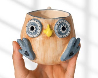 May include: A ceramic owl-shaped mug with a light brown body, blue and white eyes, a yellow beak, and blue wing-like handles. The mug is held in a hand against a white background.