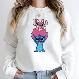 May include: White sweatshirt featuring a graphic of the characters Stitch and Angel from the animated movie Lilo & Stitch. Stitch, in blue, is holding up Angel, who is pink. The design is centered on the front of the sweatshirt.