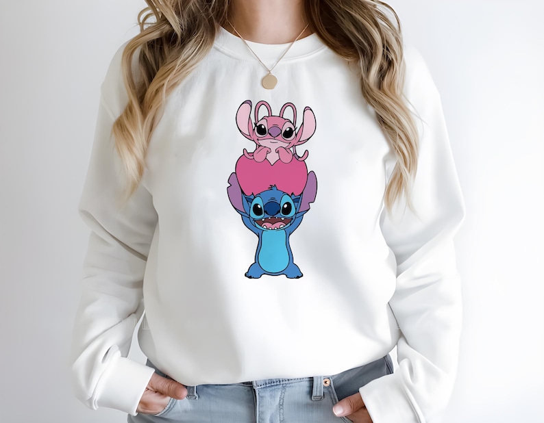 May include: White sweatshirt featuring a graphic of the characters Stitch and Angel from the animated movie Lilo & Stitch. Stitch, in blue, is holding up Angel, who is pink. The design is centered on the front of the sweatshirt.
