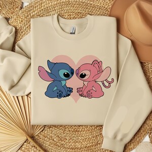 May include: A cream-colored sweatshirt featuring a cartoon illustration of two characters, one blue and one pink, facing each other in front of a pink heart. The sweatshirt has long sleeves and a crew neck.