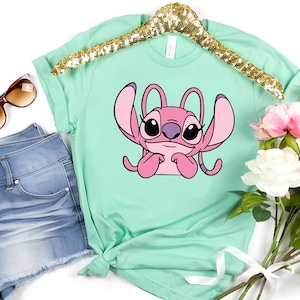 May include: A mint green t-shirt featuring a pink cartoon character with large ears and a purple nose. The shirt is displayed on a gold sequined hanger, next to a pair of blue denim shorts, sunglasses, and a bouquet of flowers.