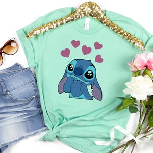 May include: Mint green t-shirt featuring a cartoon character with large eyes and blue fur, surrounded by pink hearts. The shirt is displayed on a gold sequined hanger, next to denim shorts, sunglasses, and a bouquet of flowers.