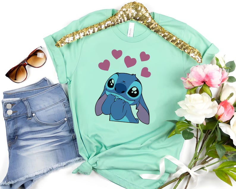 May include: Mint green t-shirt featuring a cartoon character with large eyes and blue fur, surrounded by pink hearts. The shirt is displayed on a gold sequined hanger, next to denim shorts, sunglasses, and a bouquet of flowers.