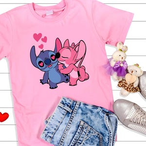 May include: Pink t-shirt featuring a graphic of two cartoon characters, one blue and one pink, embracing with hearts above them. The shirt is paired with denim shorts and silver sneakers. Two plush toys and two red heart-shaped decorations are also visible.