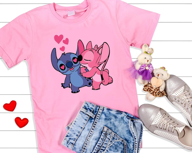 May include: Pink t-shirt featuring a graphic of two cartoon characters, one blue and one pink, embracing with hearts above them. The shirt is paired with denim shorts and silver sneakers. Two plush toys and two red heart-shaped decorations are also visible.