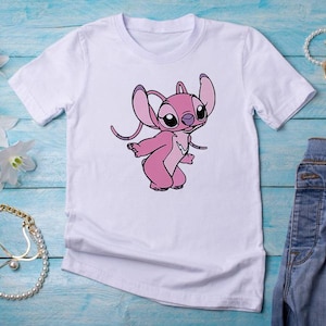 May include: White t-shirt featuring a pink cartoon character with large ears and a playful pose. The graphic is centered on the shirt. The shirt is paired with blue jeans and accessories on a blue wooden surface.