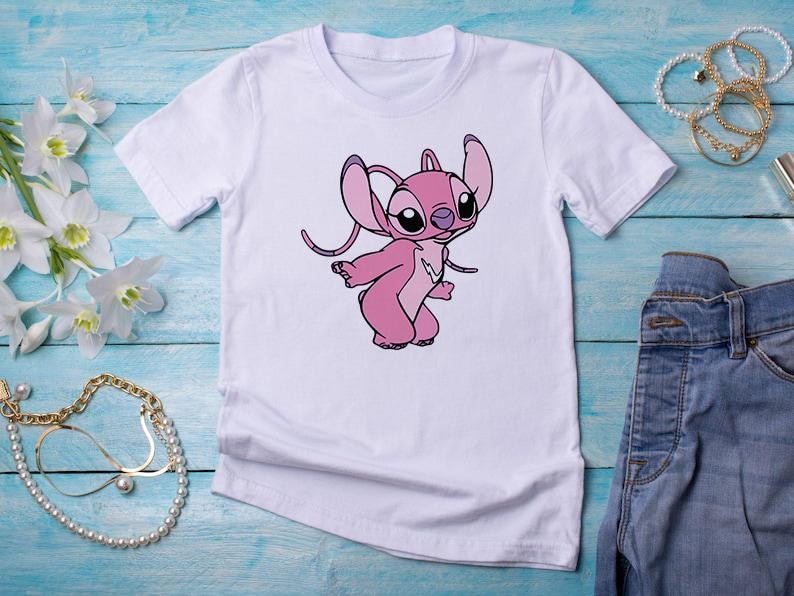 May include: White t-shirt featuring a pink cartoon character with large ears and a playful pose. The graphic is centered on the shirt. The shirt is paired with blue jeans and accessories on a blue wooden surface.
