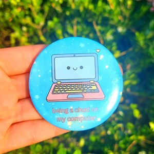 May include: A circular pinback button featuring a light blue background with white stars and a cartoon laptop with a smiling face. The text on the button reads, being a chud on my computer. The design includes a pink laptop base and yellow keys.