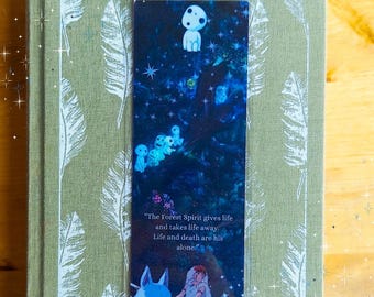 May include: A rectangular bookmark featuring a dark blue forest scene with small white spirits, a blue wolf, and a person. The text reads: The Forest Spirit gives life and takes life away. Life and death are his alone. The bookmark rests on a sage green book.