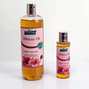Op de afbeelding: Twee flessen Hibiscusolie. De grotere fles is lang en slank, terwijl de kleinere fles korter en breder is. Beide flessen hebben zilveren doppen en roze etiketten met de tekst "Hibiscus Oil" en een illustratie van een hibiscusbloem.