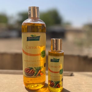 Op de afbeelding: Twee flessen wortelolie worden getoond. De grotere fles heeft een gouden dop en een etiket met de woorden "Carrot Oil" en een wortelafbeelding. De kleinere fles heeft een vergelijkbaar ontwerp. De olie heeft een gouden kleur.