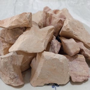 K&ouml;nnte beinhalten: Ein Haufen roher Tonbrocken in verschiedenen Gr&ouml;&szlig;en und Formen. Der Ton hat eine hellbraune Farbe mit rosa und roten Farbt&ouml;nen. Die raue Textur und die unregelm&auml;&szlig;igen Formen deuten auf ein nat&uuml;rliches, unverarbeitetes Material hin, m&ouml;glicherweise f&uuml;r Bastel- oder Kosmetikzwecke.