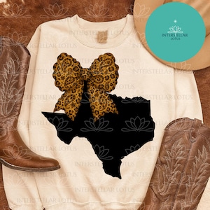 May include: Cream-colored sweatshirt featuring a black Texas outline with a leopard print bow. The design is centered on the front of the shirt. The words "INTERSTELLAR LOTUS" are printed on the shirt. Brown cowboy boots are on either side.