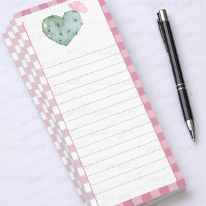 May include: A stack of lined notepads with a pink gingham border. The top sheet features a watercolor cactus heart and a small pink flower. A black pen rests on the right side of the notepad.