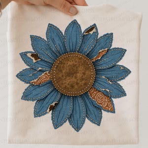 May include: A cream-colored t-shirt featuring a large, appliqued sunflower. The sunflower has blue denim petals with distressed edges and a brown center. The design is stitched onto the shirt, creating a textured effect. The text "INTERSTELLAR LOTUS" is printed on the shirt.