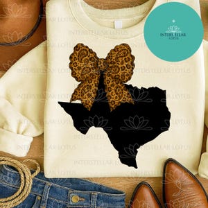 May include: Cream-colored sweatshirt featuring a black Texas outline with a leopard print bow. The design includes the text "INTERSTELLAR LOTUS" and a turquoise circle with a logo. The sweatshirt is paired with jeans and cowboy boots.