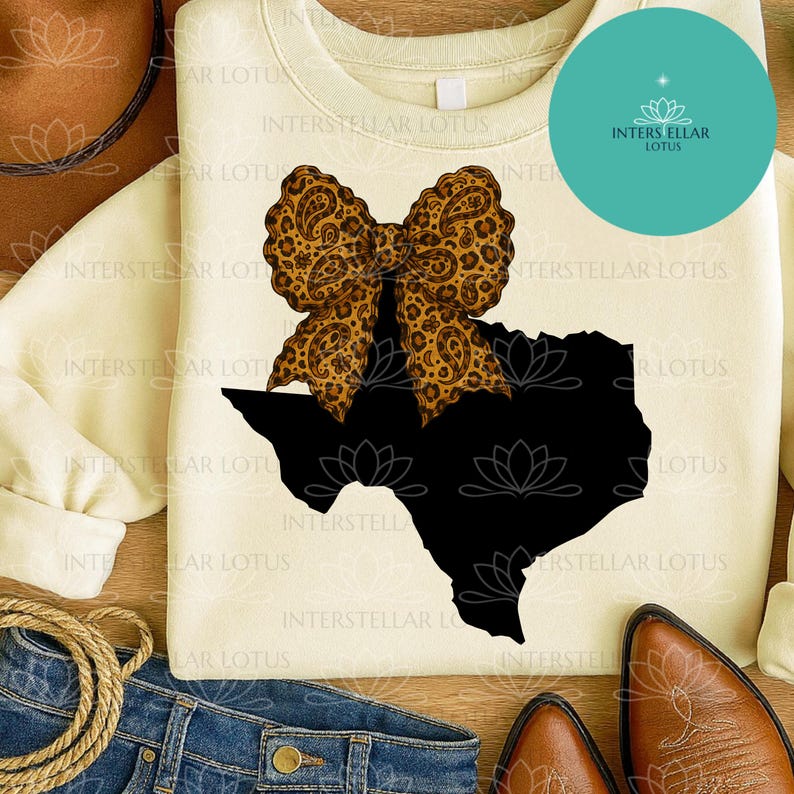 May include: Cream-colored sweatshirt featuring a black Texas outline with a leopard print bow. The design includes the text "INTERSTELLAR LOTUS" and a turquoise circle with a logo. The sweatshirt is paired with jeans and cowboy boots.