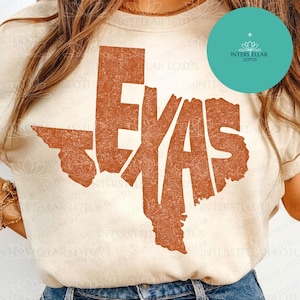 May include: A cream-colored short-sleeve t-shirt featuring a large, distressed, burnt-orange graphic of the state of Texas. The word TEXAS is integrated into the state shape in a bold, block-style font with a textured, weathered appearance.