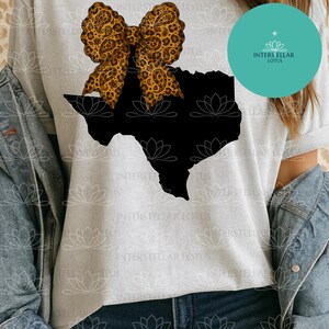 May include: A light gray t-shirt featuring a black silhouette of the state of Texas. A large bow with a brown paisley and leopard print design is positioned above the state. The shirt is part of the Interstellar Lotus collection.